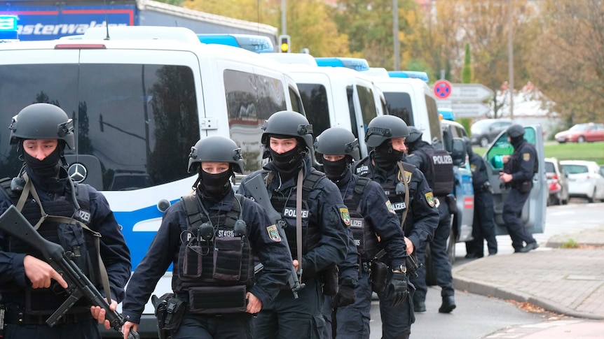 Armed police in protective gear line up with guns out next to a number of police vans