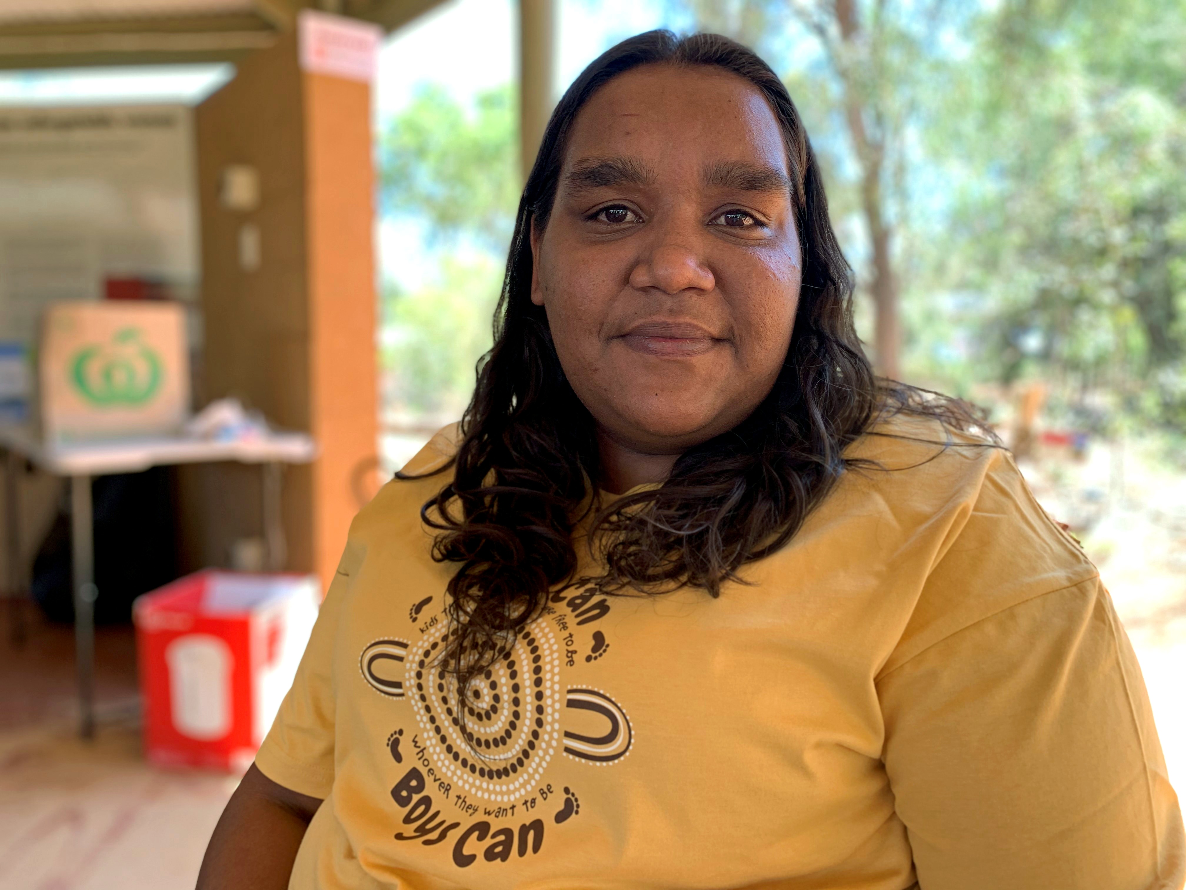 A woman wearing a shirt with an Indigenous art design on it looks at the camera with a neutral expression.