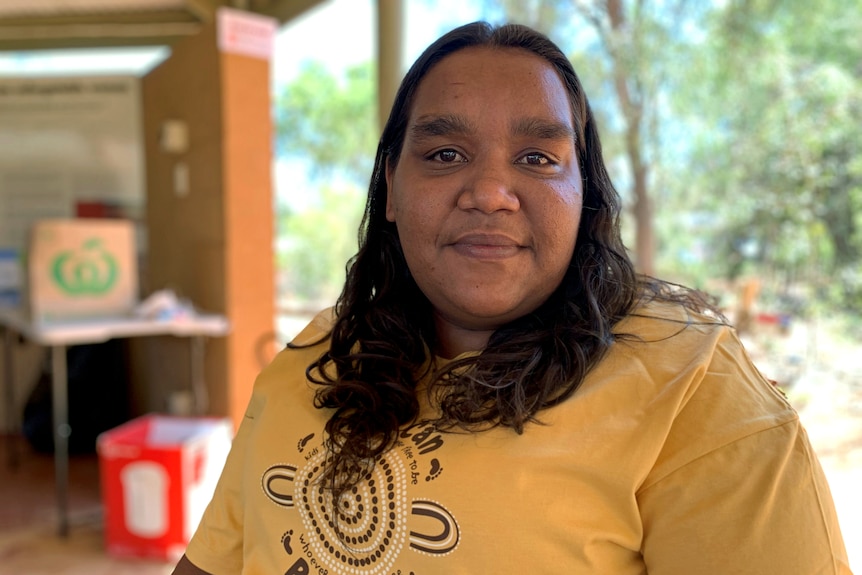 A woman wearing a shirt with an Indigenous art design on it looks at the camera with a neutral expression.