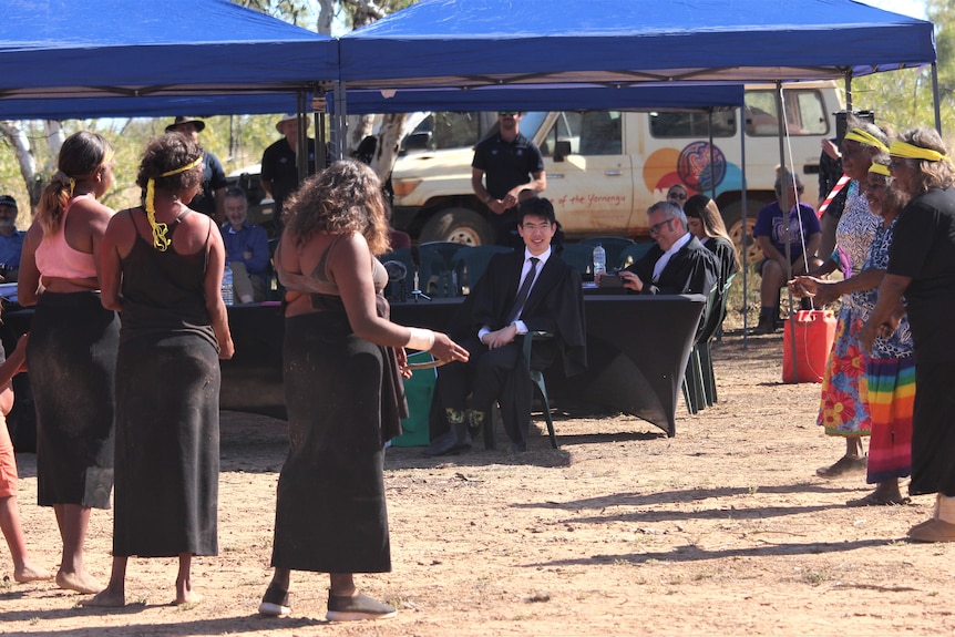 Three woman stand dancing in front of a portable gazebo filled with people in suits set up in a dry area 