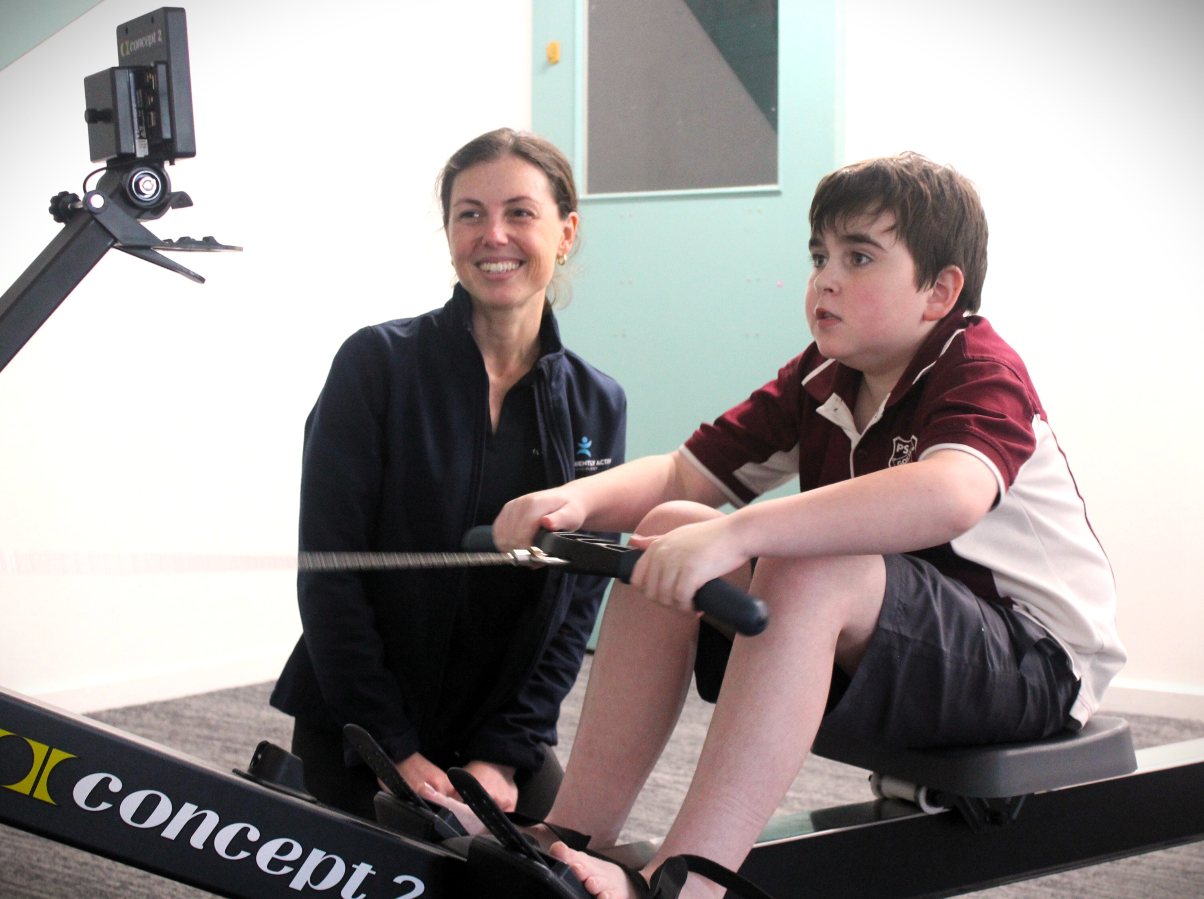 Um menino com uniforme escolar se exercitando em uma máquina de remo com uma mulher ajoelhada ao lado dele