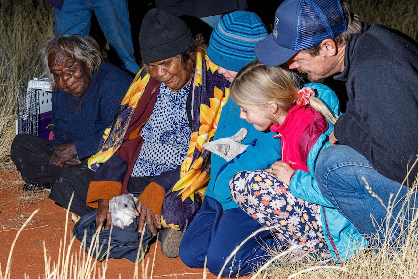 A group of people sit on the red dirt, with a bilby on the floor in front of them. They wear winter clothes.