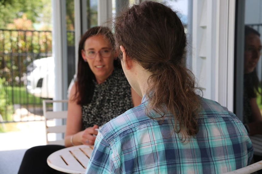 A young brunette woman in glasses smiles at ponytailed young man at table with trees behind.