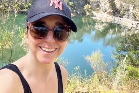 Selfie of Jill Stark smiling and wearing sunglasses and blue cap while she hikes along a river.