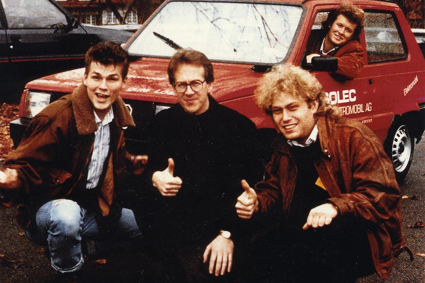 A grainy photo of four men giving the thumbs up by a small red car