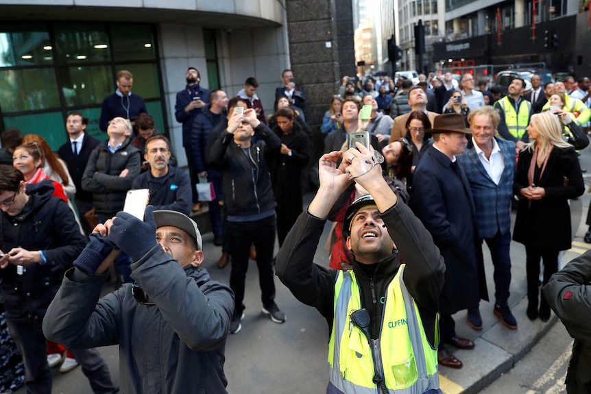 people in a city look up and hold their cameras up to take photos at something above