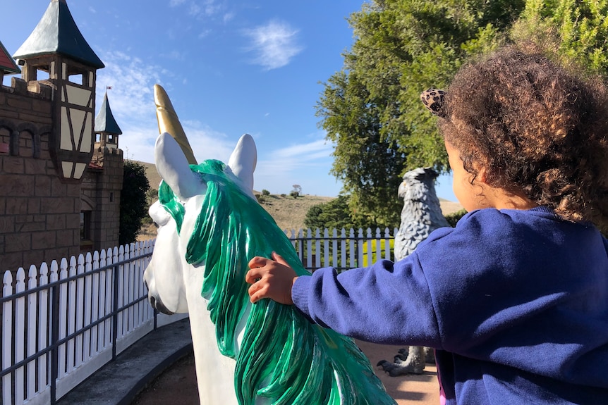 A young child with curly brown hair looks away from camera, sitting on a unicorn statue. 
