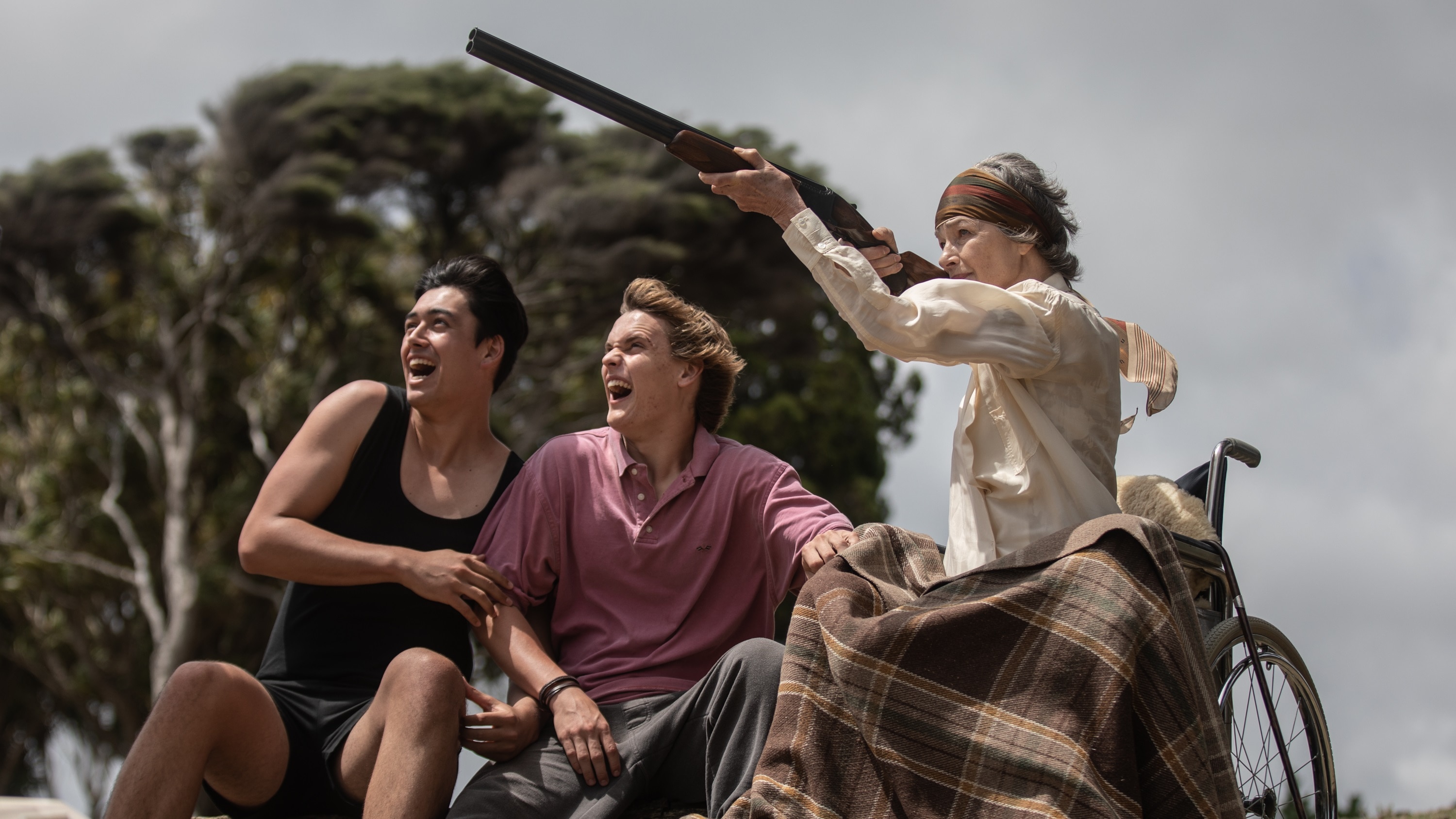 Two young men look on excitedly as they sit beside an old woman in a wheelchair who aims a gun into the air