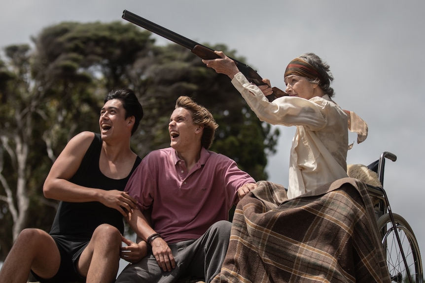 Two young men look excited as they sit next to an old woman in a wheelchair who points a gun in the air