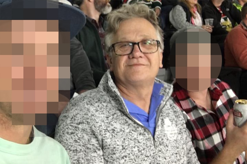 A bespectacled man sits between two younger men, apparently at a sporting match.
