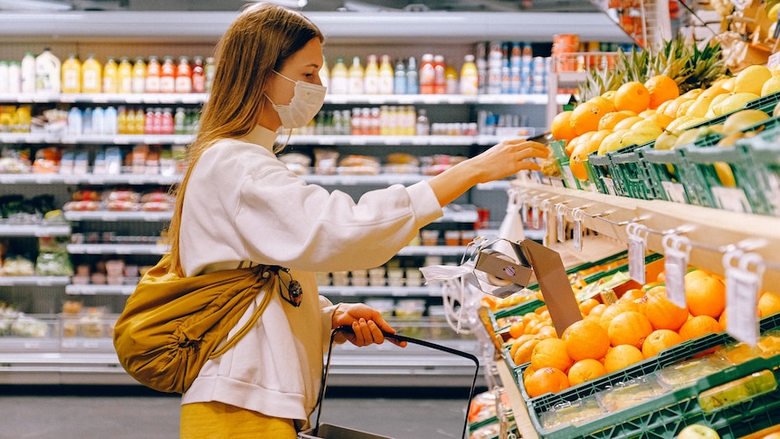 A woman wearing a face mask chooses fruit while grocery shopping with a hand basket