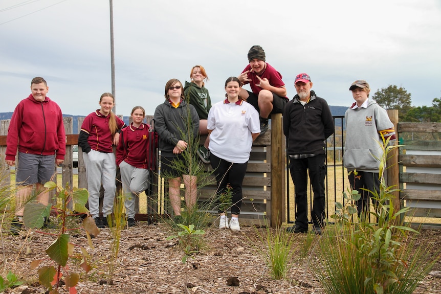 Group of smiling students stand on dirt patch with growing plants, two sit on fence, man with grey beard, red hat stands near.