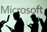 Silhouettes of laptop and mobile device users are seen next to a screen projection of Microsoft logo