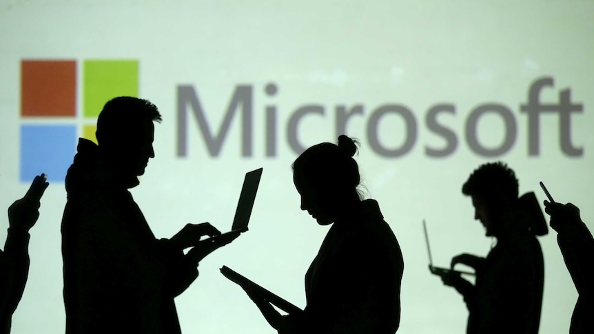 Silhouettes of laptop and mobile device users are seen next to a screen projection of Microsoft logo