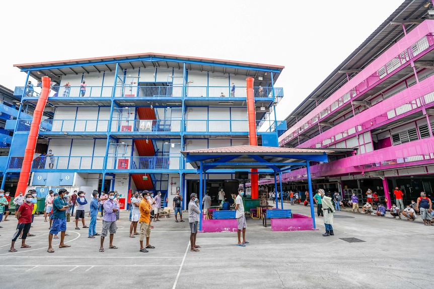 A group of men in face masks stand in a line in a court yard surrounded by brightly coloured buildings