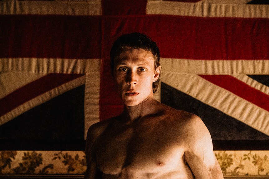 Young man barechested staring at camera with union jack flag behind him.
