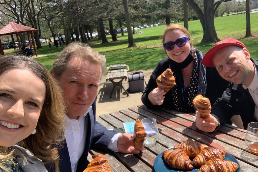 Two men and two women sitting at an outside table in gardens holding croissants for the camera.