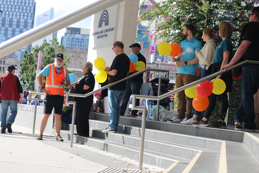 A crowd of men and woman holding brightly coloured balloons stand on steps outside the ABC in South Bank.