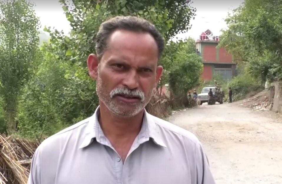 man in grey shirt speaks to the camera standing on a dirt road in pakistani-controlled kashmir