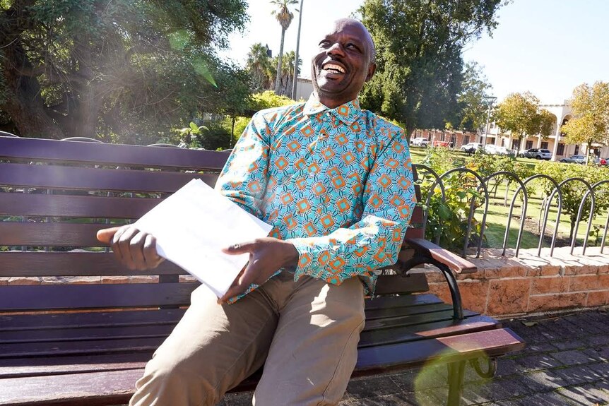 A Congolese man in a bright patterned shirt sick back on a park bench surrounded by trees laughing.