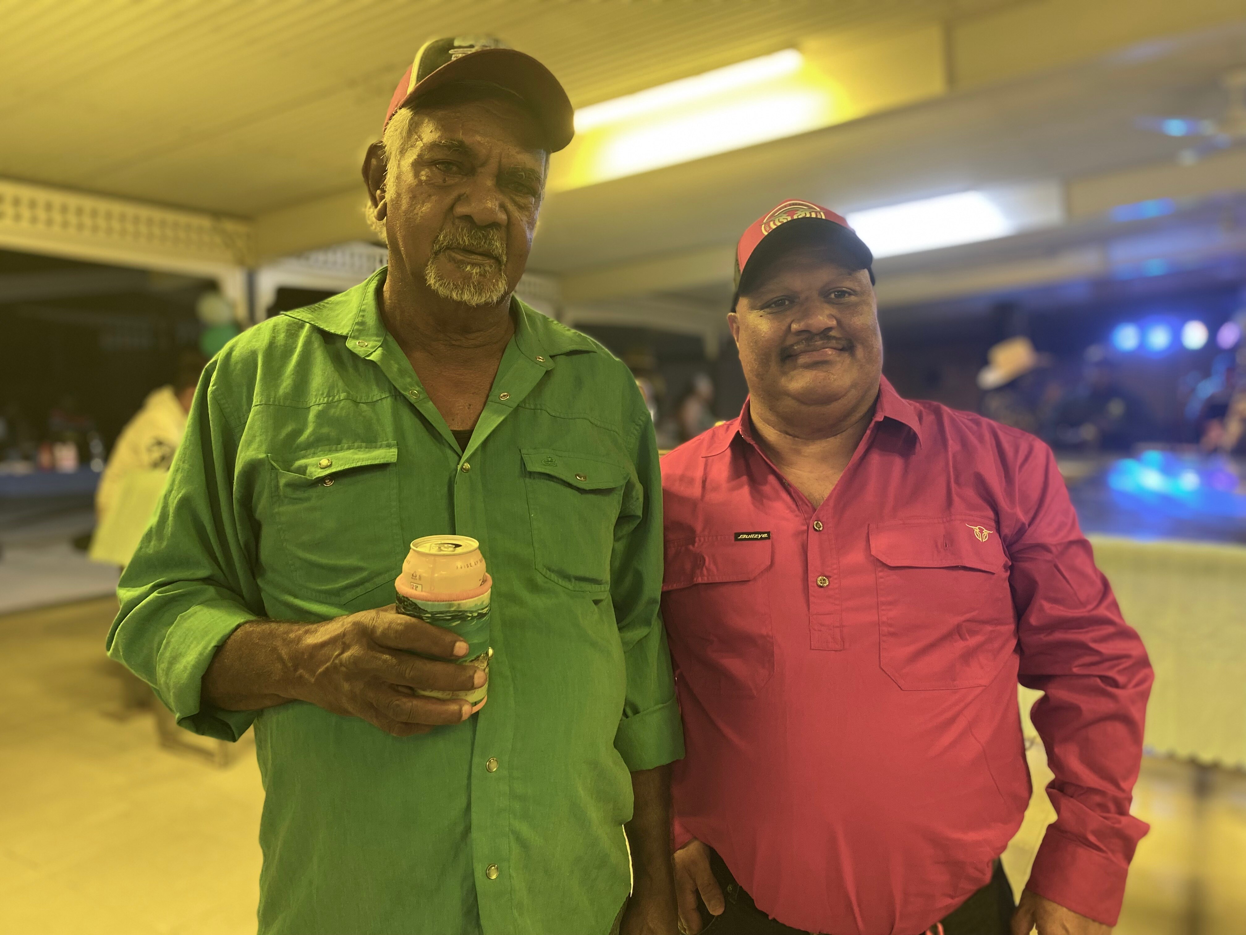 Two Indigenous men stand side by side under fluorescent lights in a bar.