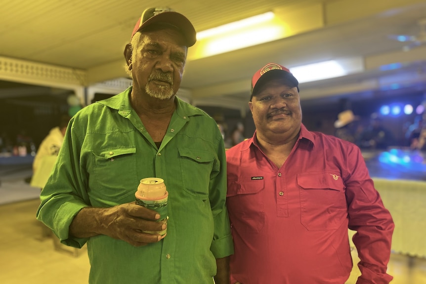 Two Indigenous men stand side by side under fluorescent lights in a bar.