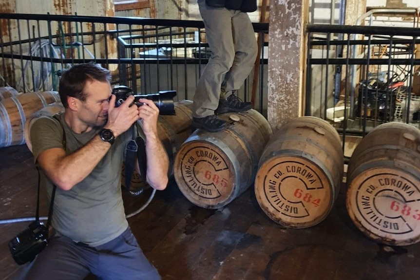 A man in a khaki T-shirt kneels on the ground on set with a camera to his eye and wine barrels stacked behind him.