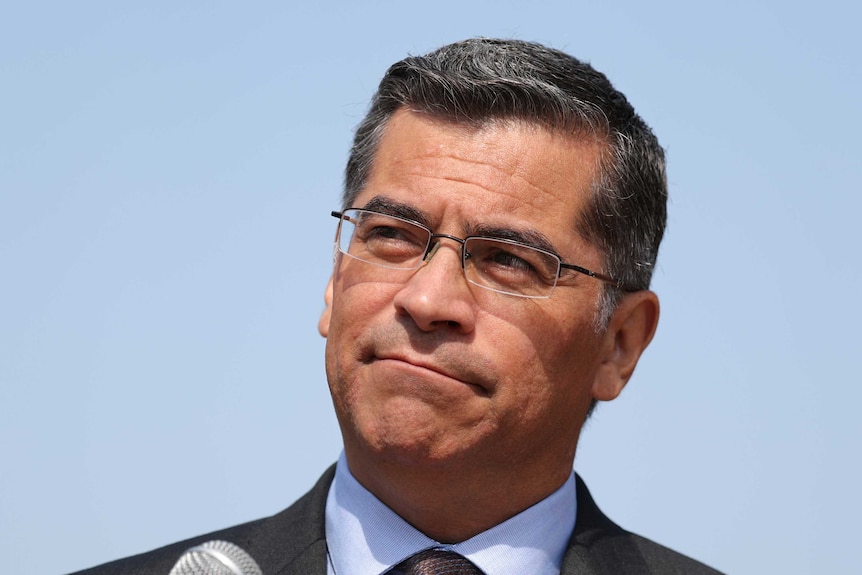 California Attorney General Xavier Becerra wears glasses standing in front of blue sky and looks off-camera