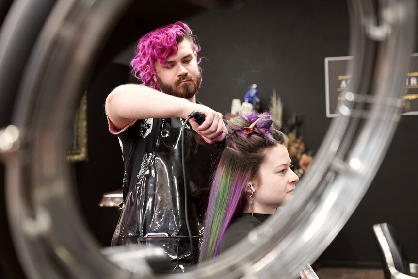 A hairdresser with bright pink hair is working on a client with purple and green hair inside a salon 