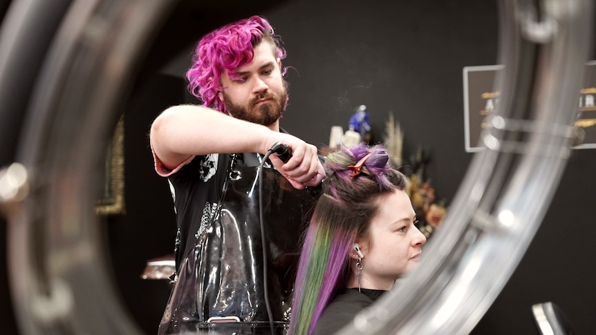 A hairdresser with bright pink hair is working on a client with purple and green hair inside a salon.