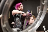 A hairdresser with bright pink hair is working on a client with purple and green hair inside a salon.