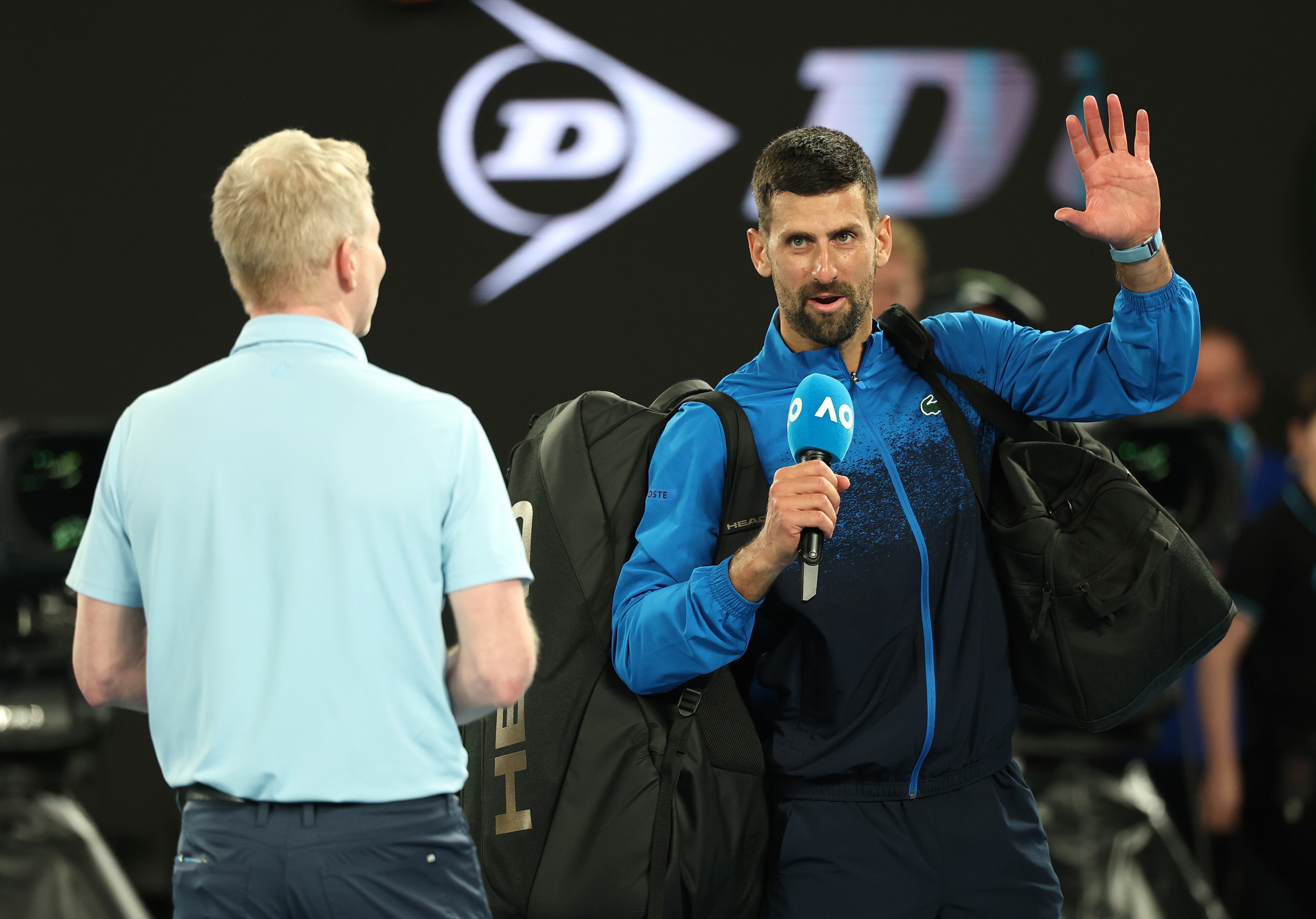 Novak Djokovic speaking into a microphone, waving to the crowd in appreciation