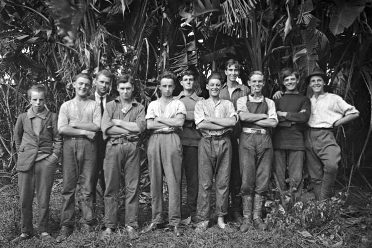     A group of teenage boys in casual trousers and shirts stand in front of a tree in 1922.
