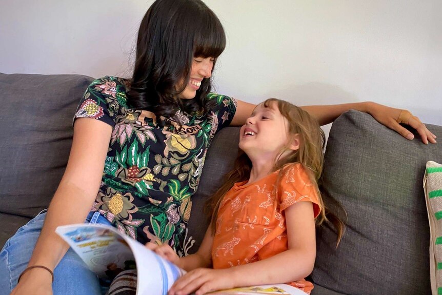 A mother and daughter sitting on a grey couch laughing and reading a book.