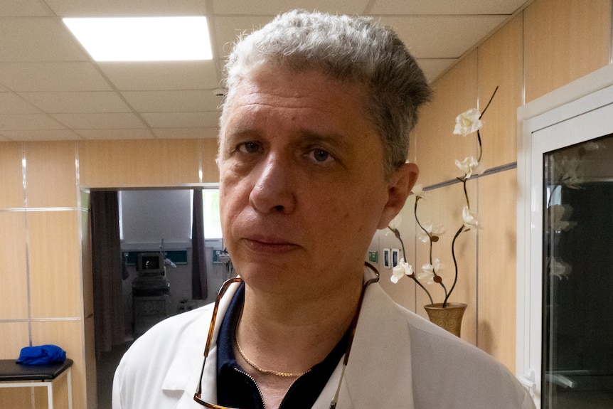 A male doctor in in blue scrubs stands in front of equipment in a hospital room.