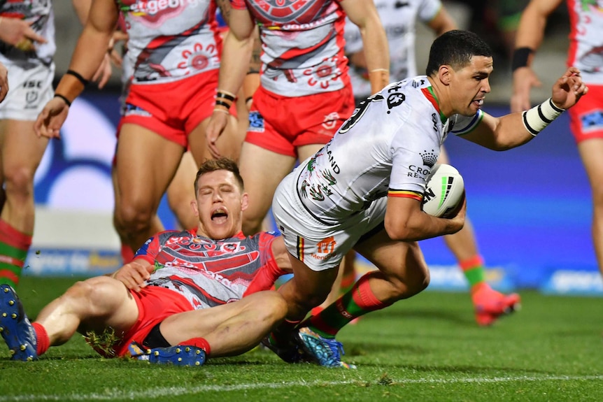 An Indigenous NRL player dives over, holding the ball in his right hand with his other arm raised.