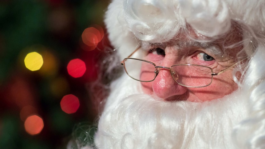 Santa peers over his glasses as Christmas lights twinkle behind him.