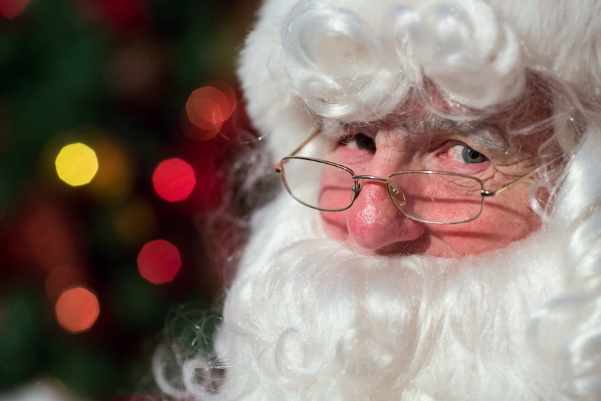 Santa peers over his glasses as Christmas lights twinkle behind him.
