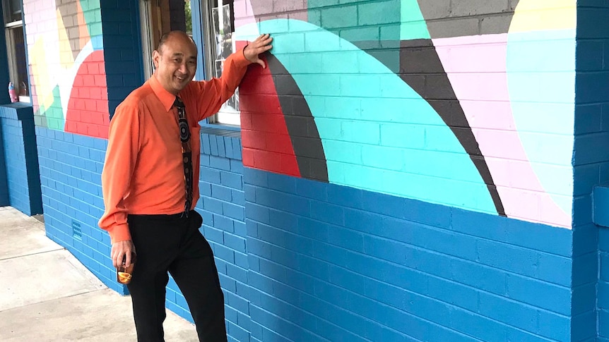 A middle-aged Chinese-Australian man smiles as he touches a pastel-coloured mural on a blue wall.