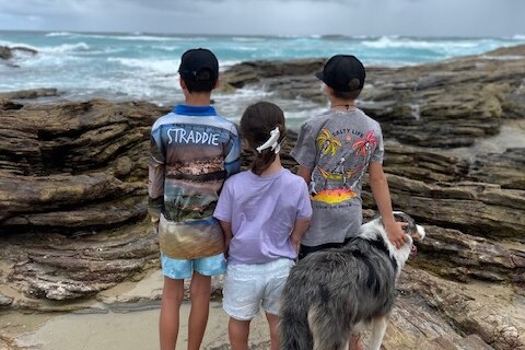 a photo of three siblings looking out over the ocean at the beach. one of the brothers is patting a large dog