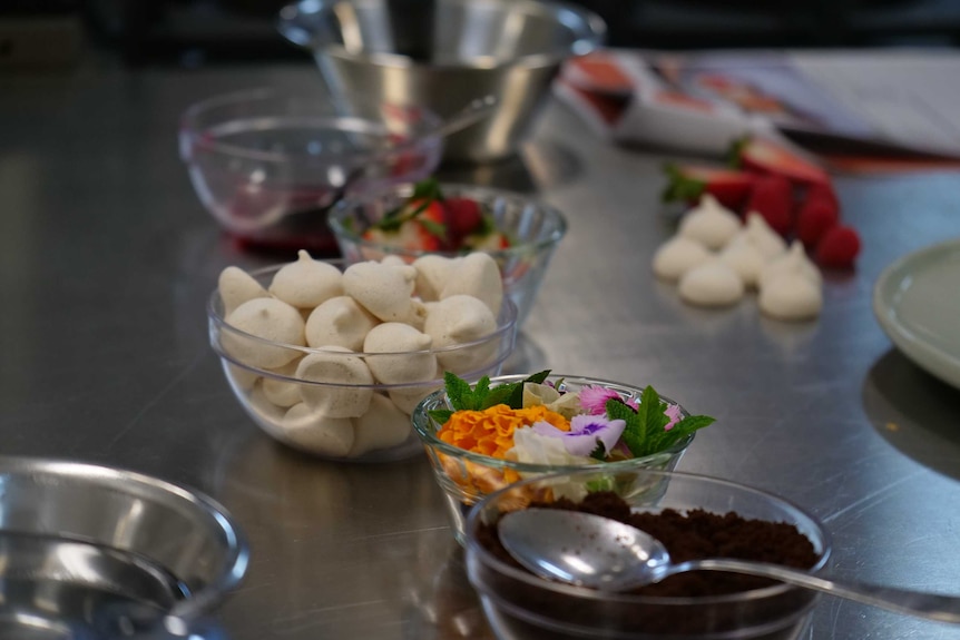 Clear bowls containing decorative flowers and mint leaves, and small meringues and strawberries.