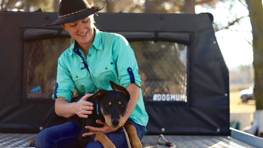 A woman with an akubra sitting in the back tray of a ute hugging her farm dog who's on her lap.