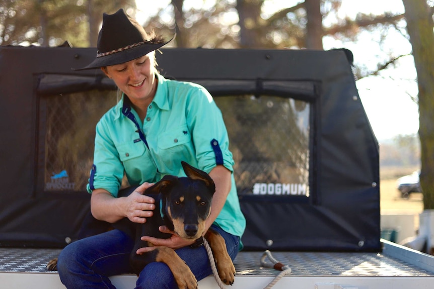 A woman with an akubra sitting in the back tray of a ute hugging her farm dog who's on her lap.