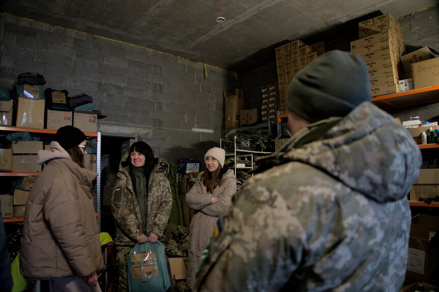 Women in military gear and jackets stand in a group in a warehouse. 