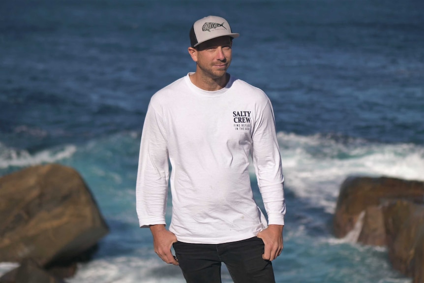 A man wearing a long sleeve t-shirt and a cap stands in front of a coastal scene.