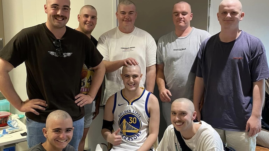 A group of young men with shaved heads standing around a young man in a chair in a hospital room 