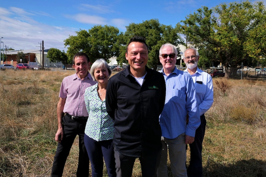 A grey-haired woman and a four men stand on a vacant lot, all smiling widely.