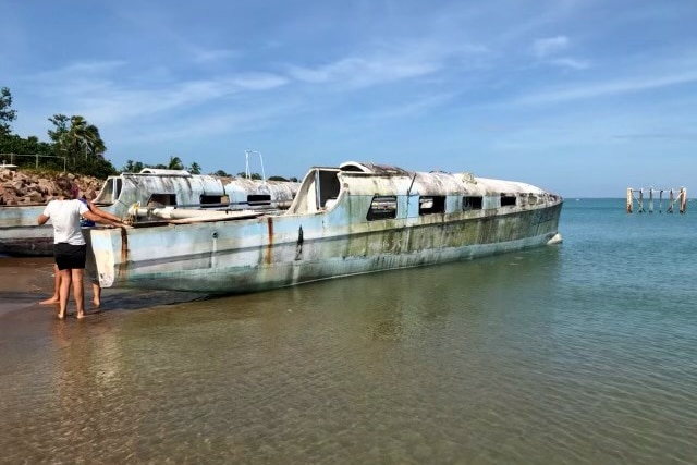 Old and rundown white catamaran sits in shallow waters