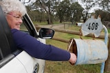 A woman with grey hair and glasses sits in car and reaches out to check the postbox.