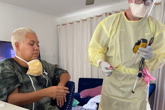 A woman sits on chair while a doctor dressed head to toe in plastic conducts checks.